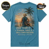 Blue t-shirt featuring a cowboy with a flag-patterned guitar on a beach at sunset with patriotic text printed on the back.!