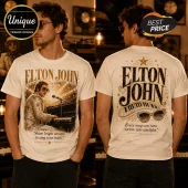 Cream-colored t-shirt featuring Elton John playing a piano on the front and his name with star motifs on the back.!