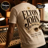 White crewneck t-shirt featuring metallic gold Elton John text, piano keys, star motifs, and rhinestone-style sunglasses.!