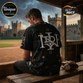Man wearing black Harry Potter themed baseball jersey with house initials sitting on wooden bench by a baseball field.!