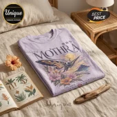 Lavender t-shirt featuring a Mothra graphic with floral accents and Japanese text laid on a beige bedspread.!
