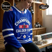 Toronto Marlies blue hockey jersey featuring 2026 Calder Cup Champions graphic, white stripe accents, and maple leaf icon.!