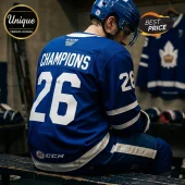 Hockey player wearing a blue Toronto-style jersey with number 26 and Champions text sitting on a bench in a locker room.!