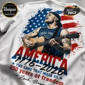 White t-shirt graphic featuring Zach Bryan playing acoustic guitar over a red, white, and blue American flag splatter print.!