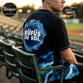Black baseball jersey with blue crystal graphic and Rufus Du Sol text worn by a person sitting in stadium seating.!