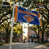 New York Knicks 2026 NBA Champions blue and orange flag flying on a metal pole against a blurred outdoor park background.!